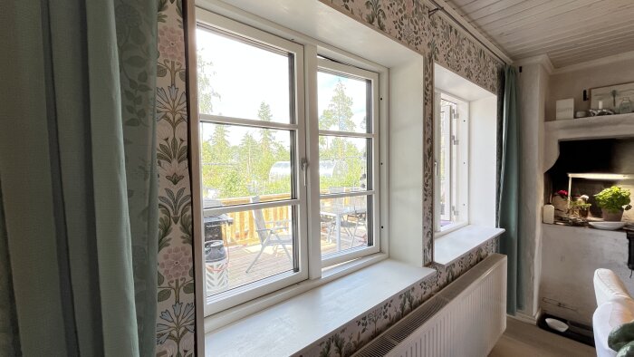 Inomhusbild på ett fönster med utsikt mot en uteplats. Fönstersalningen är vitmålad och rummet har mönstrad tapet och gröna gardiner.