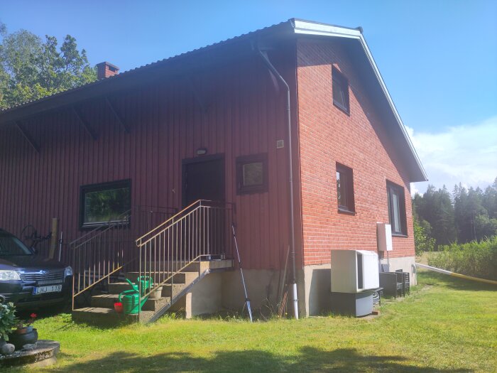 Husfasad med rödmålad panel som möter tegelfasad i husets hörn, trappa med ledstång på vänster sida, gröna växter i förgrunden.