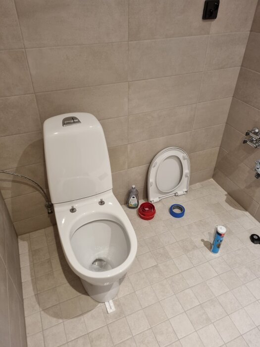 Vit toalettstol monterad i ett kaklat badrum med silikon och verktyg på golvet. Toalock står bredvid.