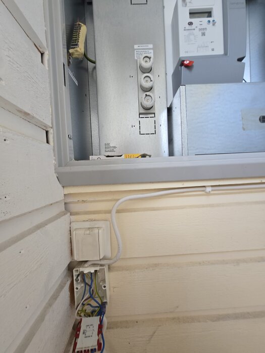 Elcentral med mätarskåp och säkringar monterad på vägg; under denna finns en vit strömbrytare med elledningar och kopplingsdosa.