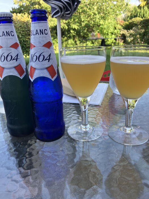 Två flaskor och två glas med öl står på ett glasbord i en trädgård med grönska i bakgrunden.