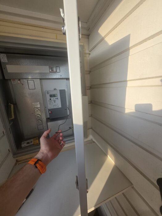 En hand sträcker sig mot en elcentral med kablar, synlig genom en öppen dörr på väggen med ljus träpanelsklädsel.