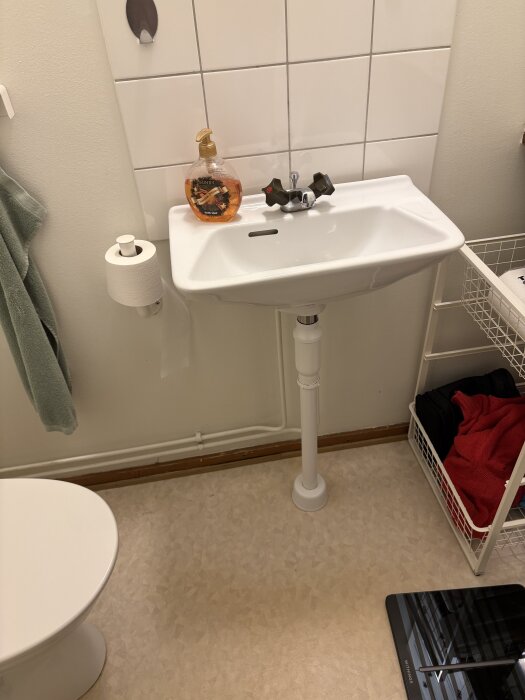 Litet badrum med vitt handfat och kran, tvålflaska ovanpå, toalett till vänster, handdukshållare med grön handduk, kaklade väggar och korgställ.