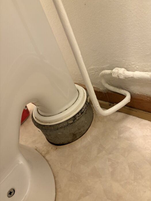 Baksida av en toalettstol med avloppsrör och en närliggande vattenledning på ett beige golv.