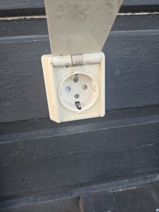 Uttag med trasigt lock och kabelspår på mörk fasad.