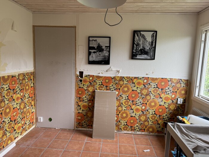 Rum med delvis gipsad dörröppning och färgglad blommig tapet, svartvita foton på väggen och klinkergolv.