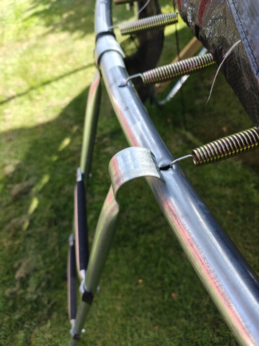 Trampolinsteg som hänger löst på en metallram med fjädrar synliga i bakgrunden, på en gräsmatta.