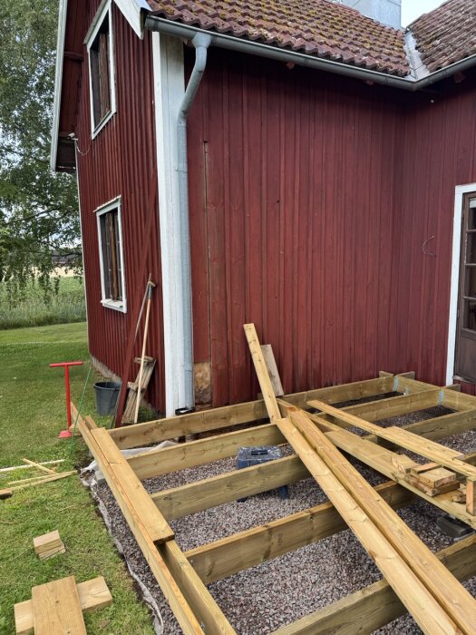 Röd träfasad med bristfällig panel vid stuprör, byggnation av altan. Träbjälkar och plankor på mark täckt av grus.