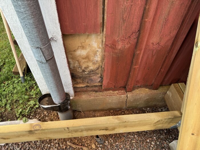 Sprucken väggpanel på husgrund vid stuprör, med fuktskador och rödfärgad träpanel. En del av altanbygge syns.
