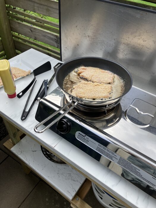 Stekt schnitzel tillagas i en stekpanna på ett utekök med grill, senapsflaska på bänken.