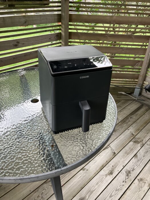 Svart airfryer på ett glasbord i ett utekök med trägolv och omgiven av spaljé.