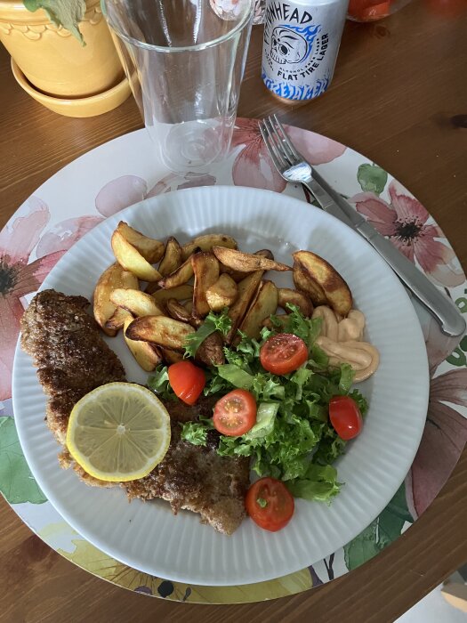 Stekt schnitzel med citron, potatisklyftor, sallad, tomat och gurkskivor på en tallrik. Hamburgerdressing som sås. En kanna och ölburk i bakgrunden.