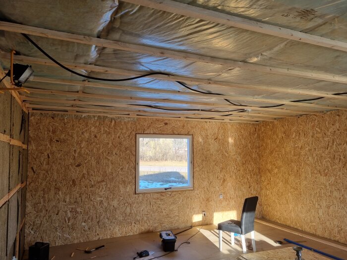 Rum med OSB-väggar, isolerat tak och ett fönster; fåtölj och verktyg syns på golvet, indikerar pågående byggprojekt.