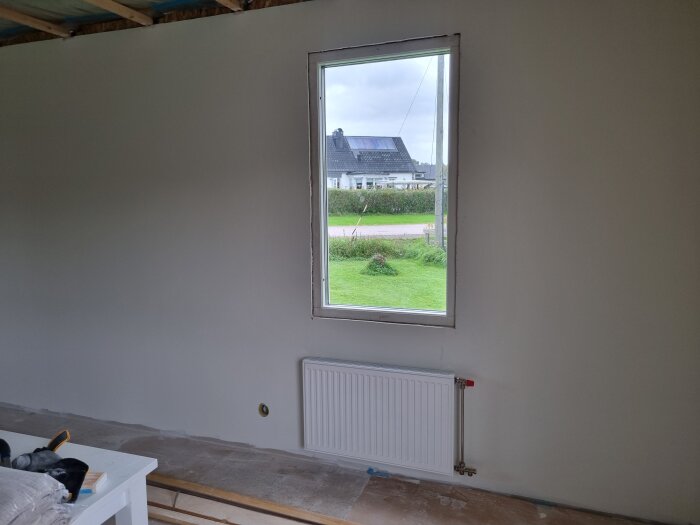 Vitt rum med ett fönster, utsikt mot ett hus och grönska. Omonterad sockel, målade väggar och en vit radiator syns.