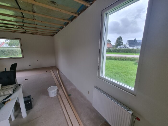 Nyrenoverat rum med ett fönster som vetter mot en trädgård, synliga träreglar i taket, och ett element under fönstret.