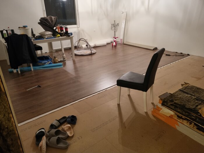 Rum under renovering med laminatgolv, stol, bord med verktyg, barnvagnsdyna och leksakscykel. Diverse objekt på golvet inklusive skor.