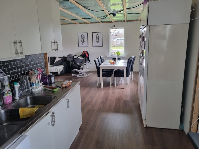 Köksvy med vit inredning, svart kakel, matbord med stolar, soffhörna, barnvagn och tak under konstruktion.