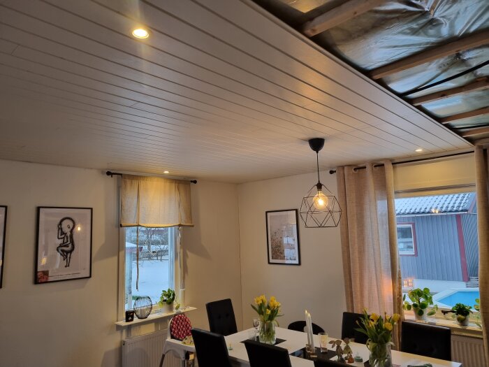 Renoverad matsal med vita väggar, takpaneler och fönster med gardiner. Taklampa i geometrisk design, bord dekorerat med vaser och ljus.