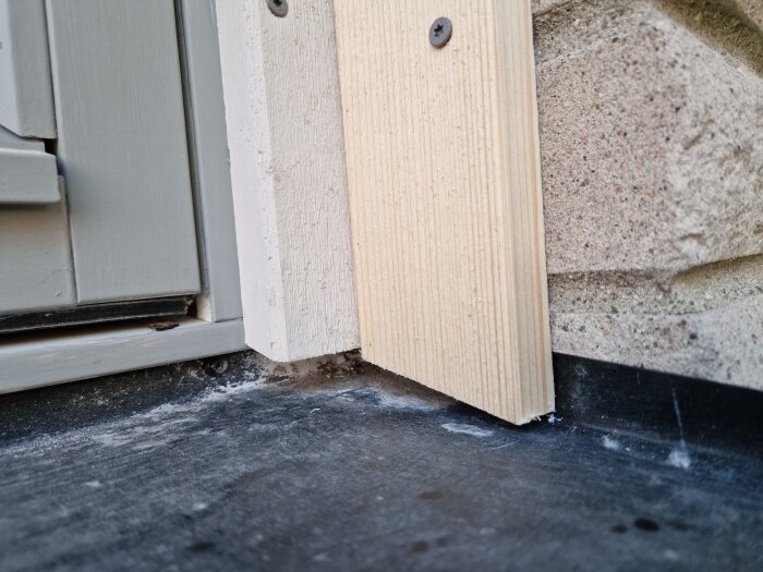 Träpanel monterad vid fönsterram med svart botten och skruvade fast. Bakgrund av stenfasad.