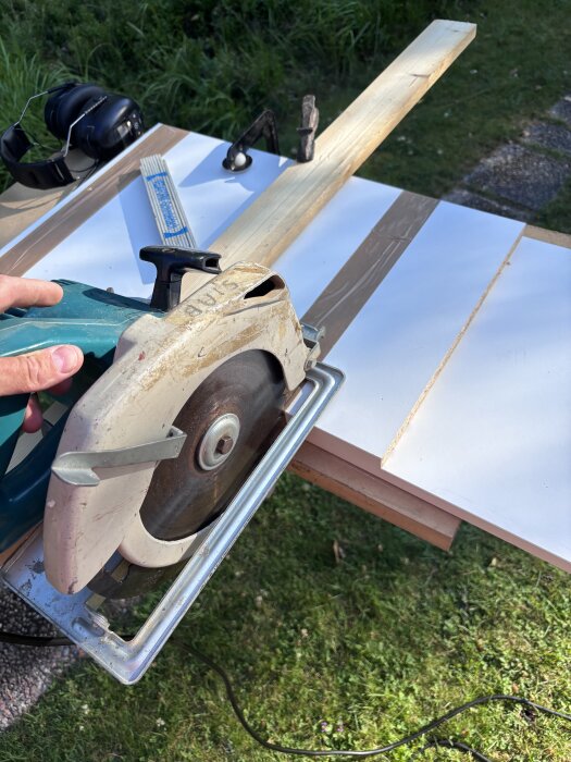 Cirkelsåg används för att kapa vit skiva med trälist ovanpå, hörselskydd och måttband syns på arbetsbänken utomhus.
