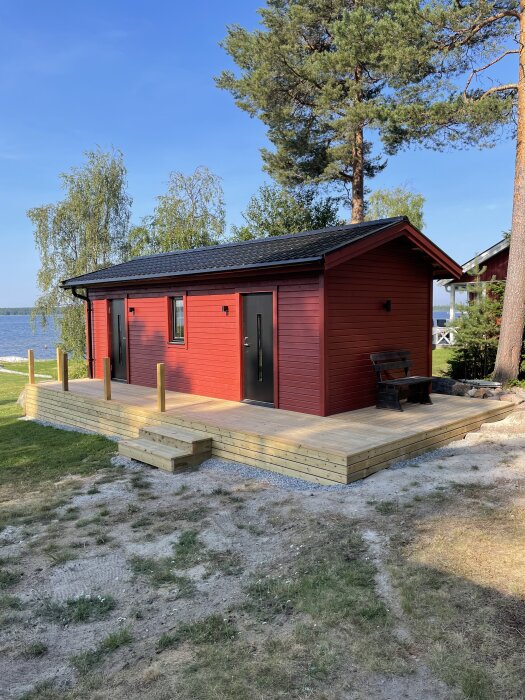 Röd stuga med nybyggd altan utan överliggare på räcket, liten trappa på kortsidan. Träd och sjö i bakgrunden.