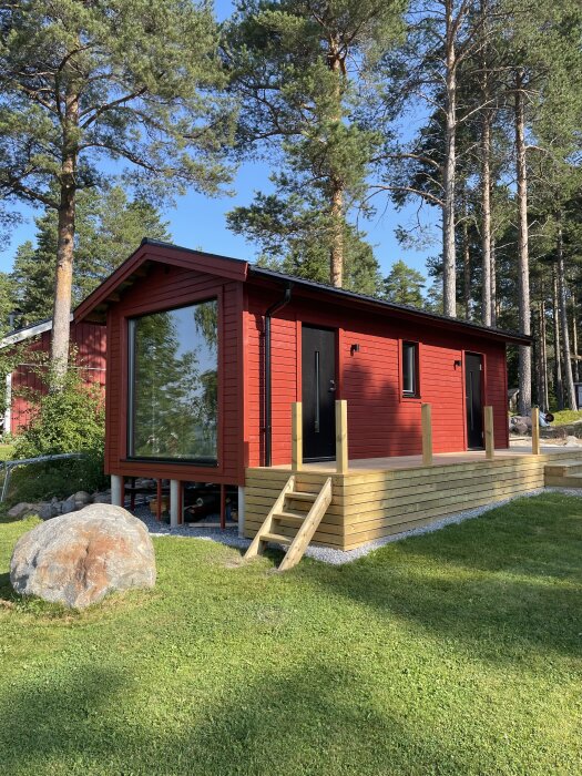 Röd byggnad med nästan färdig altan, trappa på kortsidan och skog i bakgrunden. Soligt väder.