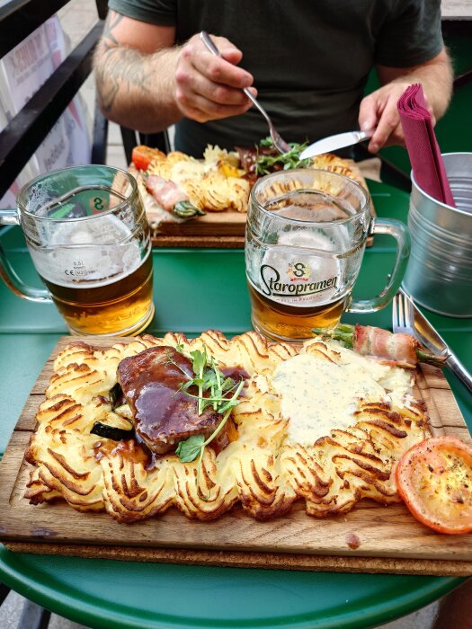 Tallrik med plankstek, potatismos, sås, baconinlindad sparris och tomatskiva, serverad med två ölglas på en grön utomhusbord.