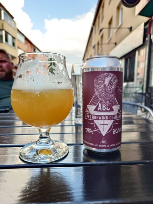 Ett glas med gyllene öl bredvid en burk från Apex Brewing Company på ett utomhusbord, med en suddig bakgrund av husfasader.