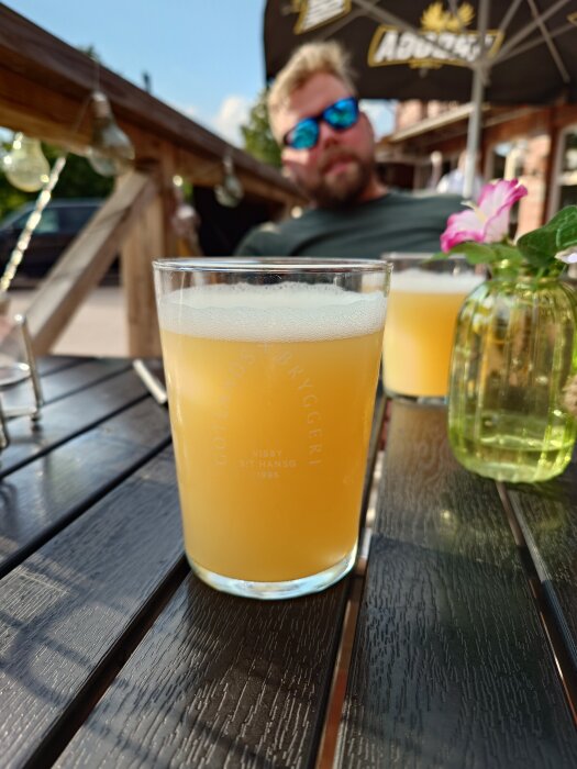 Ett glas ljus öl på ett bord utomhus med en blomma i bakgrunden och en person i solglasögon.