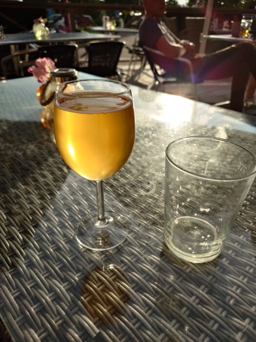Ett glas med gyllene öl står på ett vävt bord i solljus, med en tomt glas bredvid och suddig bakgrund av folk på uteservering.