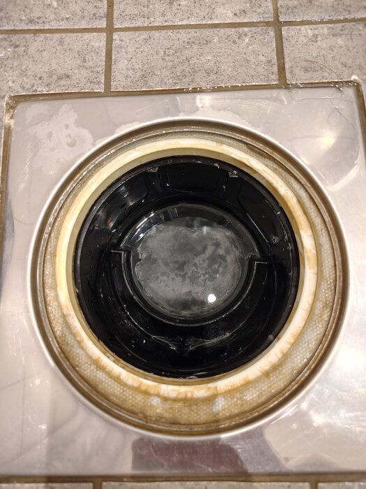 Svart rörlig del i botten av en golvbrunn, omgiven av en vit ring och metallram i ett kaklat badrumsgolv.