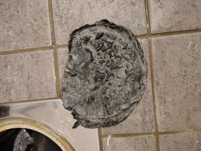 Gummiaktigt objekt funnet i botten av golvbrunn på kaklat badrumsgolv, med antydan av sedimentering eller funktion, nära avloppets öppning.