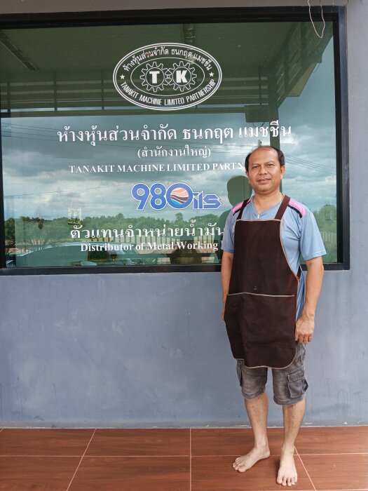 Man i förkläde står framför ett fönster till en verkstad med texten "Tanakit Machine Limited Partnership.