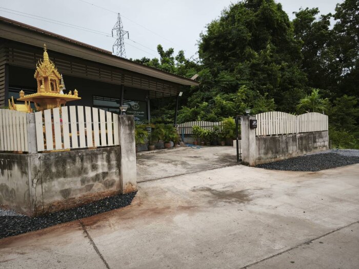 Gul byggnad i thailändsk stil bakom staket, omgiven av träd och växter. Asfalt och grus syns i förgrunden.