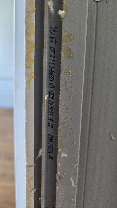 Närbild av en karm med skriven text på, omgiven av missfärgningar och intorkade rester, troligen identifierande information för fönstertillverkare.