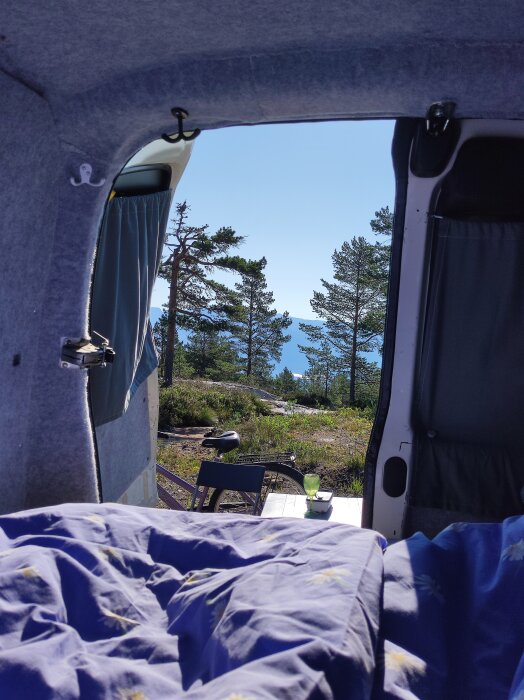 Utsikt från en campervan mot natur med träd och hav, cykel parkerad bredvid ett bord utanför.