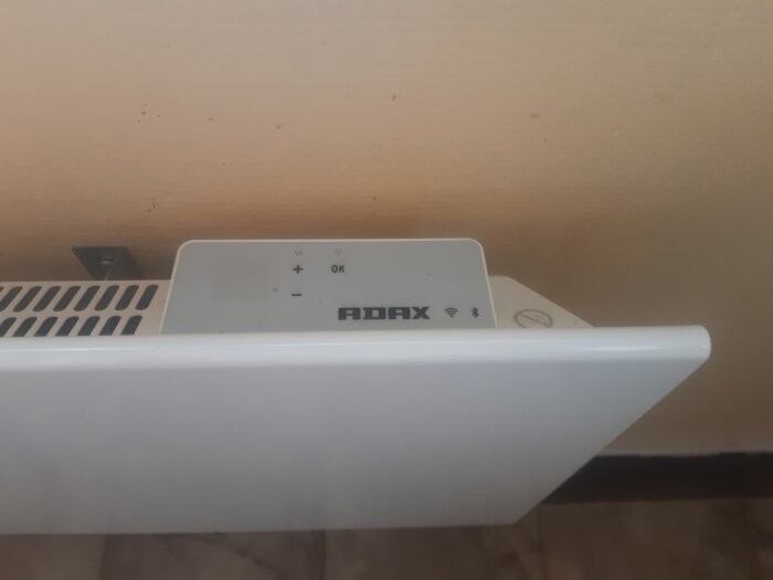 Ny, vit Adax elradiator med WiFi-funktion, styrenhet och modernt utseende, monterad på beige vägg.