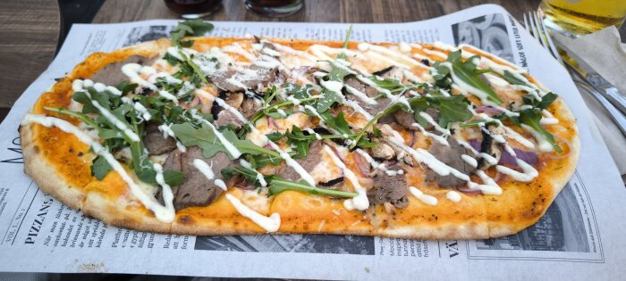 Rektangulär pizza med tomatsås, nötkött, ruccola, rödlök och smält ost, toppad med strimlad dressing och serverad på ett papper på ett bord.