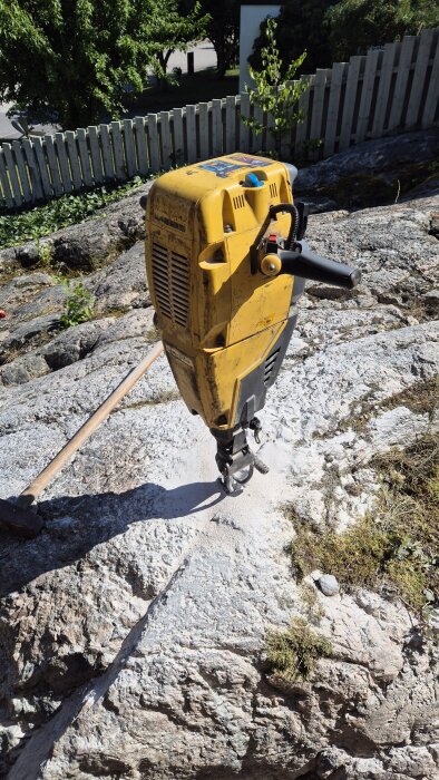 Gul borrmaskin placerad på ett stenblock utomhus, omgiven av grönska och ett vitt staket i bakgrunden.