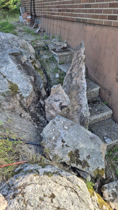 En sprucken stenformation vid en husvägg med synliga stora stenar och mossa. Gräs och verktyg kan ses runtomkring.