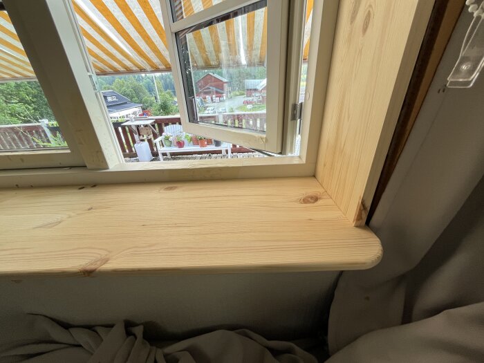 Öppet fönster med nyligen monterad och rundad fönstersalning i trä, utsikt mot terrass med växter och randig markis.