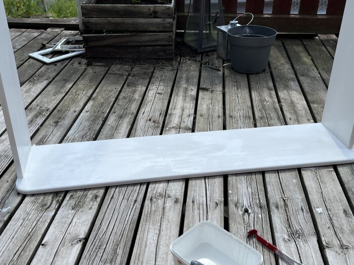 Målad fönstersalning med rundade hörn på en träveranda, omgiven av några verktyg och en vit färgburk.