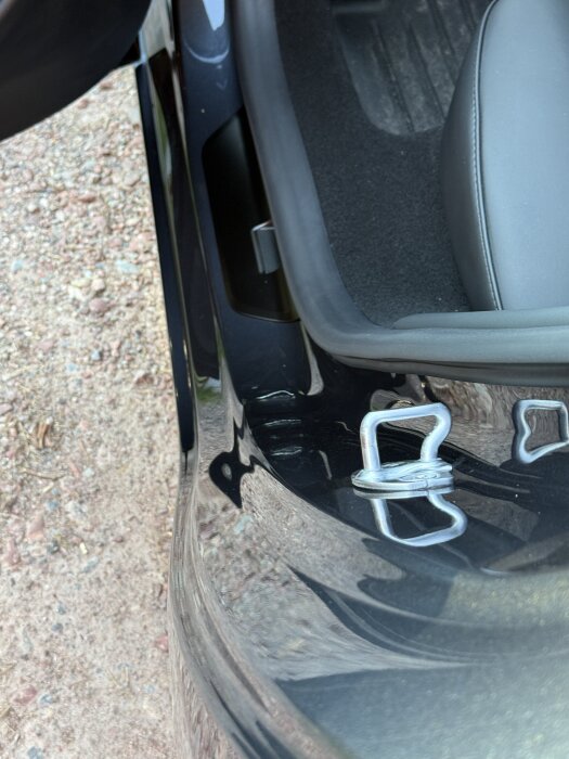 Bilens frunköppning med närbild på metallögla och del av inredningen. Utomhusmiljö med grus i bakgrunden.