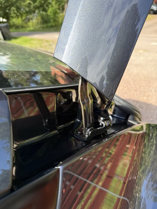 Bilden visar en detalj av en bils dörrgång med en öppnad frunk, omgiven av gröna träd och en asfaltväg i bakgrunden.