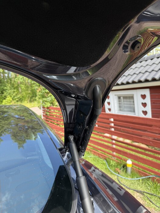 Bilens baklucka öppen, visar gångjärn och stötdämpare, med trädgård och rött staket i bakgrunden.