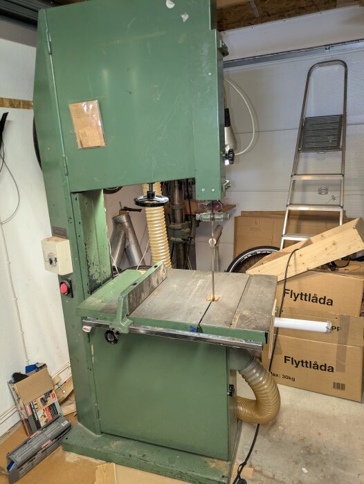 Grön bandsåg med trasig bladspänning i ett garage; flyttlådor och verktyg omkring.