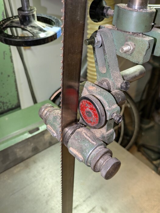 Närbild på en bandsåg med trasig bladspänning och synliga justeringsskruvar. Verktyget står på ett grönt underlag med dammig yta.