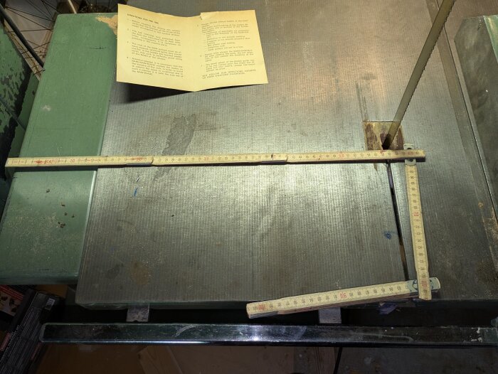 Bild av ett bandsågbord med ett måttband utlagt i vinkel för mätning. Ett pappersdokument med instruktioner ligger på bordet.