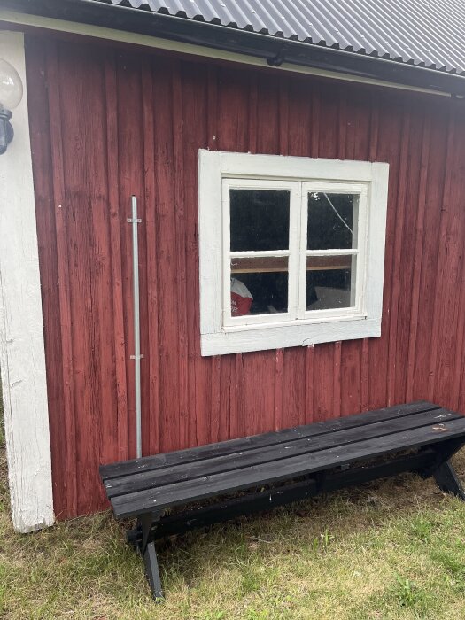 Röd vedbod med vitmålat fönster och svart bänk utanför på gräsmatta. Plåttak syns ovanför väggen.