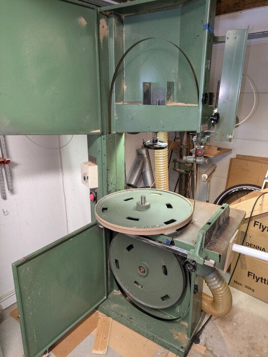 Grön bandsåg med öppna dörrar, visande blad och dubbla hjul. Maskin i verkstadsmiljö, trasig bladspänning och sågspån synliga.